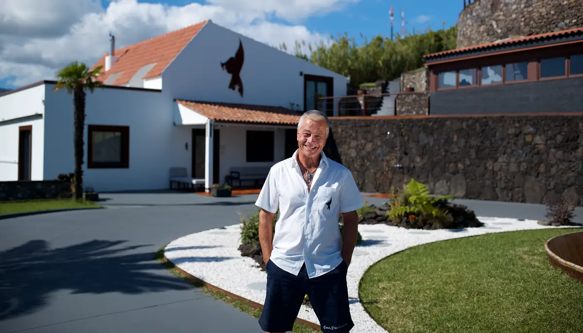 Smiling man in a white short-sleeve shirt and dark shorts stands outdoors in front of a modern white house with a stone wall and landscaped yard.